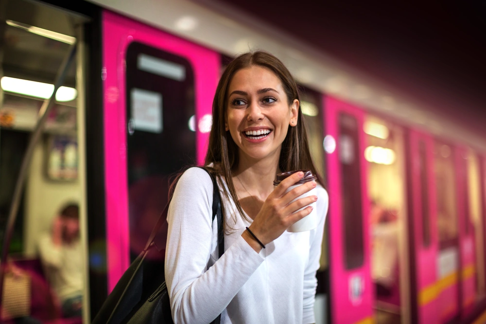 billyard_ÖPNV_KEY-Visual_300dpi_sRGB_komprimiert Lächelnde Frau mit Kaffeebecher in der Hand steigt aus einer Straßenbahn aus.